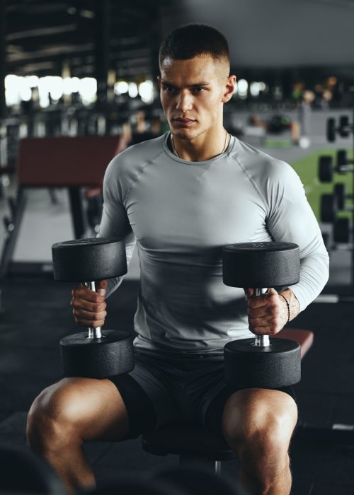 Vertical photo of a determined man preparing for his strength training workout with two heavy dumbbells in modern gym.