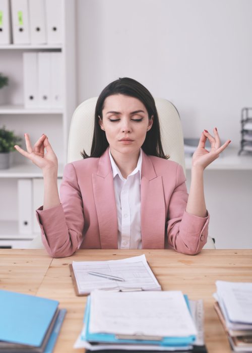 Young serious female office manager practicing yoga by desk while having break after work with financial documents or analyzing data